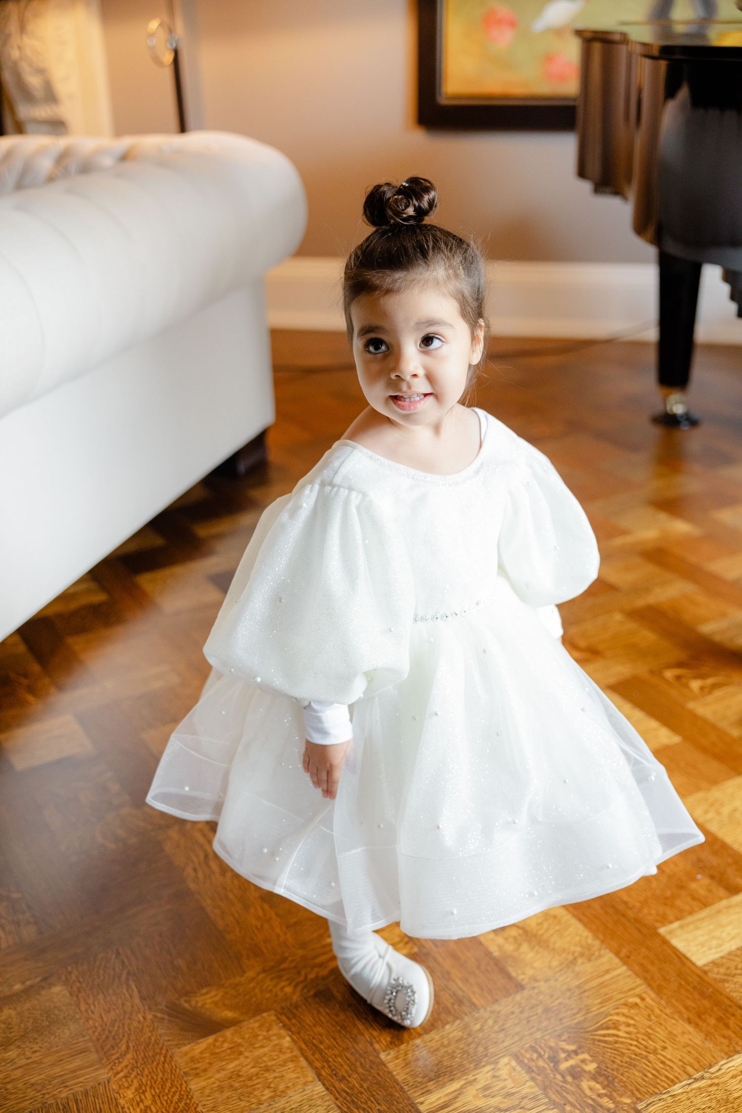 A small attendant in a white puffy-sleeved dress poses on hardwood floors.