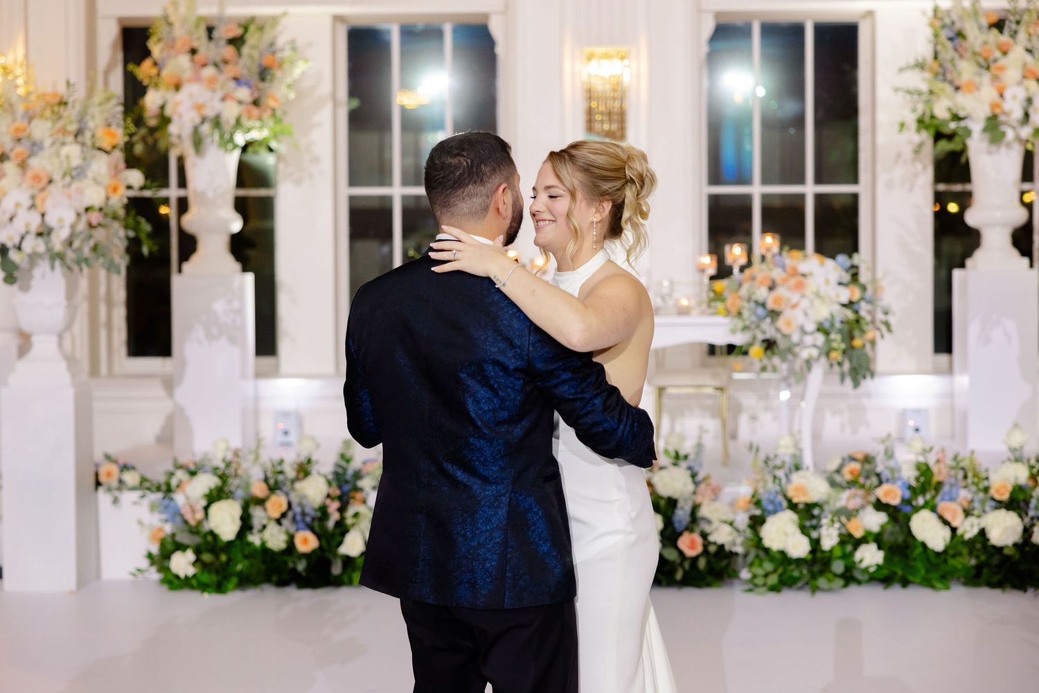 A couple shares their first dance in an elegant white ballroom decorated with pink and white floral arrangements.