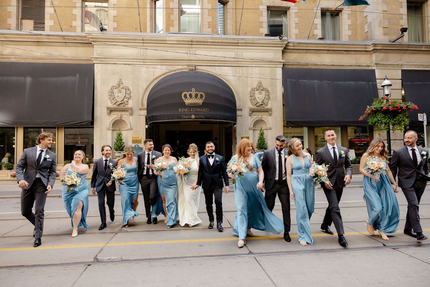 Wedding party in light blue bridesmaid dresses walks in line outside historic stone building.