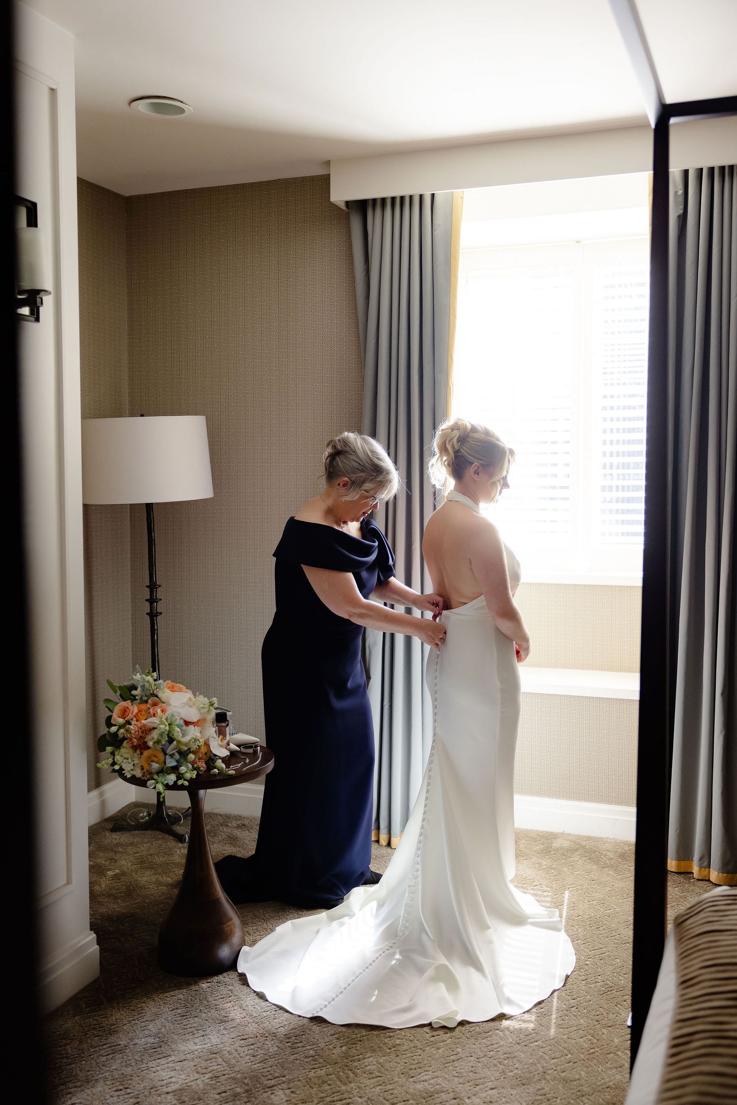Wedding dress fitting moment in a sunlit hotel room with elegant decor.