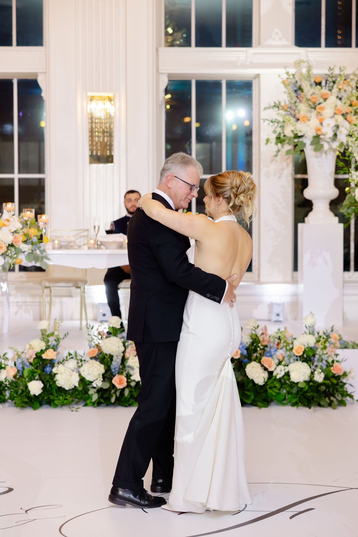 Couple dancing together in an elegant white ballroom decorated with floral arrangements and soft lighting.