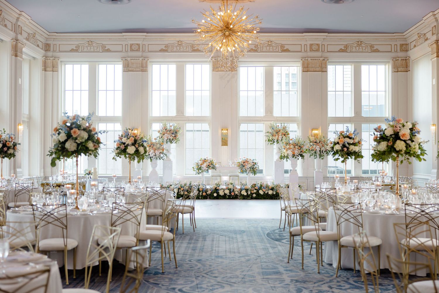 Elegant white wedding reception hall with crystal chandeliers and floral centerpieces on round tables.