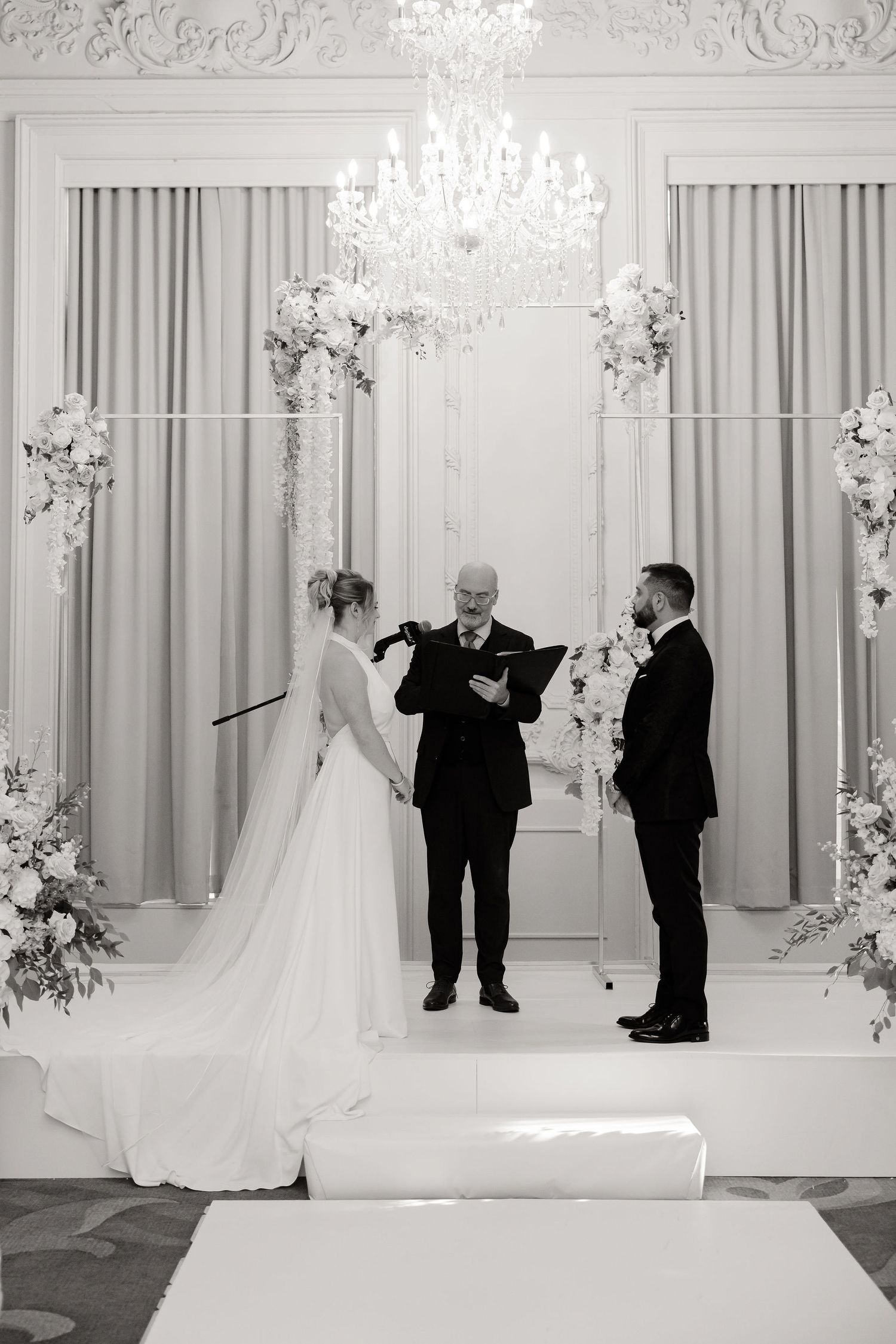Black and white photo of wedding ceremony in ornate ballroom with dramatic lighting.