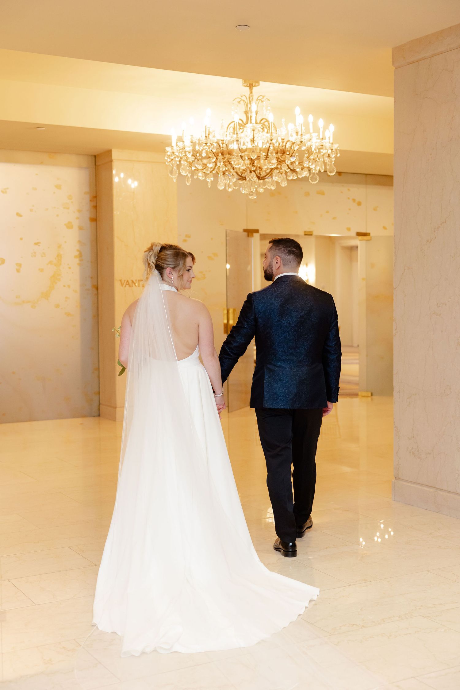 A bride and groom walk together through an elegant white ballroom with crystal chandeliers overhead.