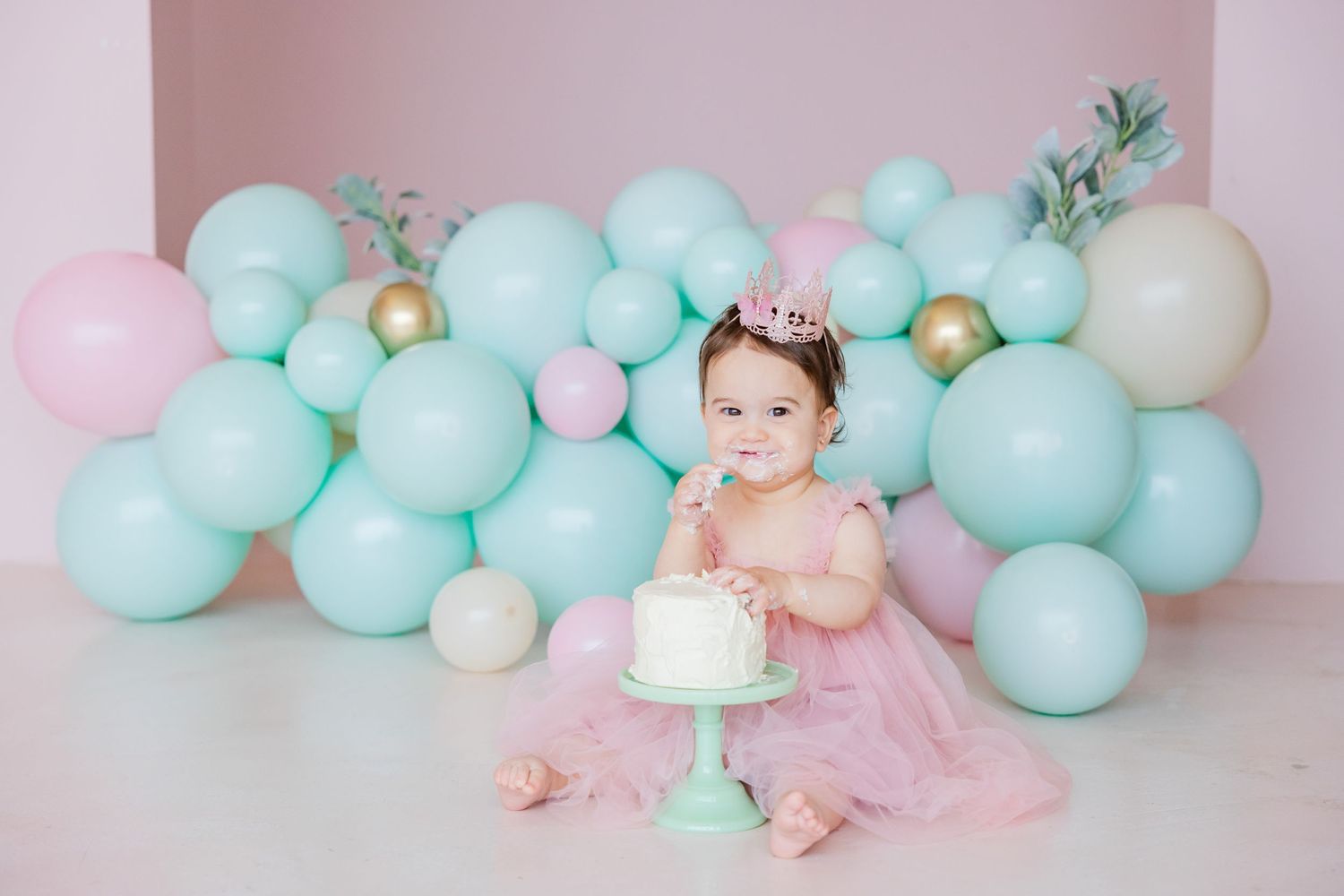 Birthday celebration setup with mint and pink balloons and a small cake on a pedestal against a pink backdrop.