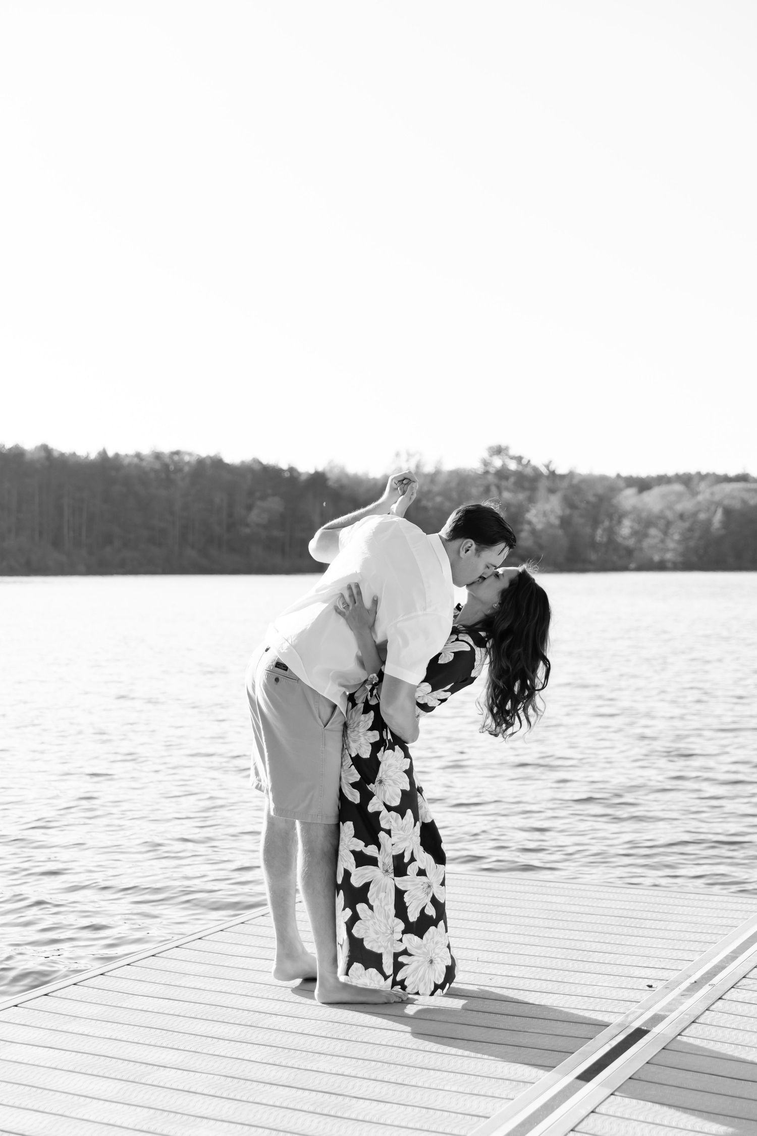 A romantic couple shares an intimate dip kiss on a wooden dock by a serene lake in black and white.