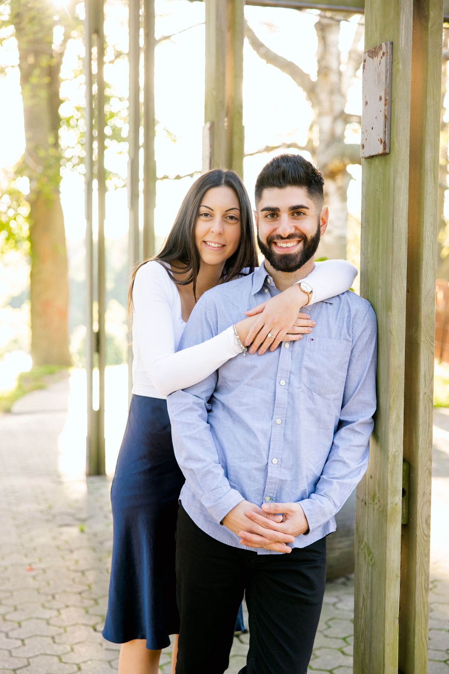 A joyful couple embracing in front of wooden pillars with natural sunlight filtering through trees.