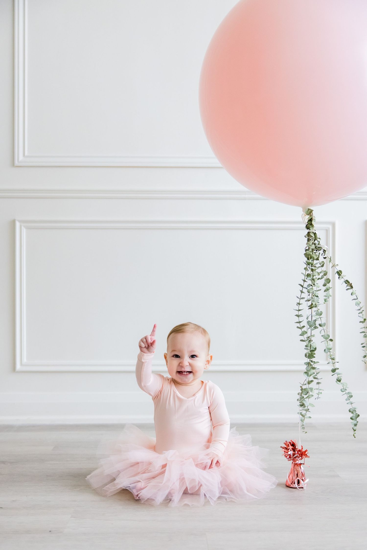 Smiling toddler in pink tutu dress points up while sitting near large pink balloon.