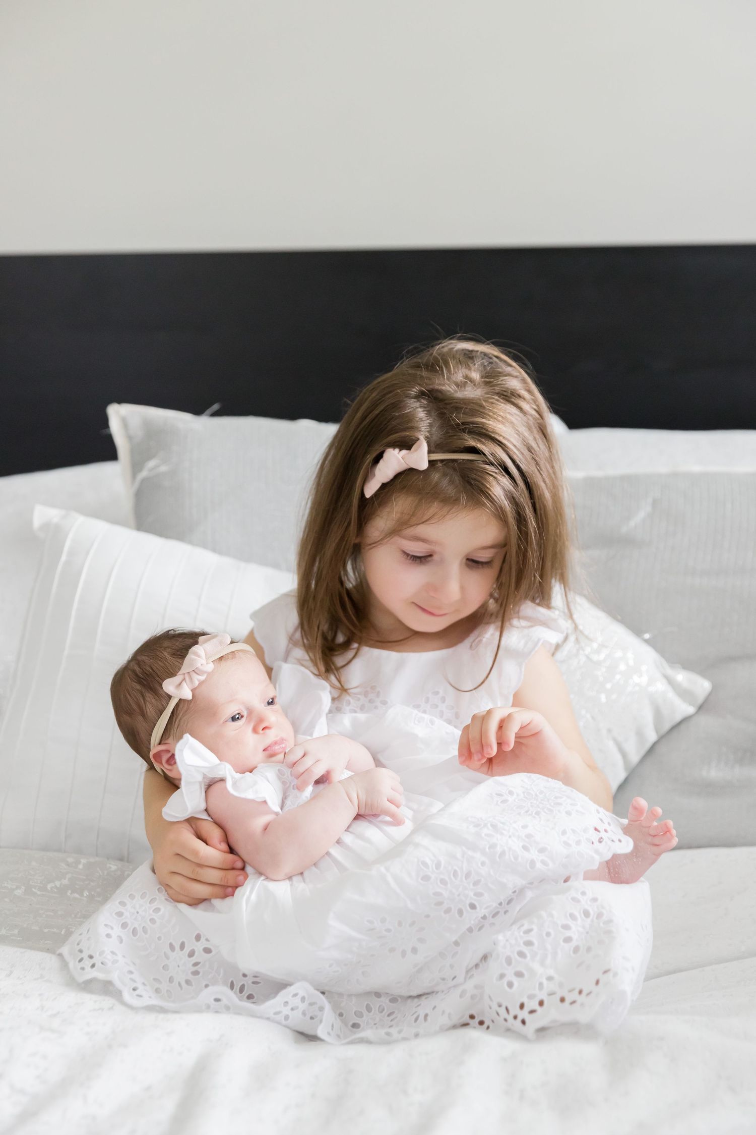 Young girl in white dress holding baby on bed with white pillows.