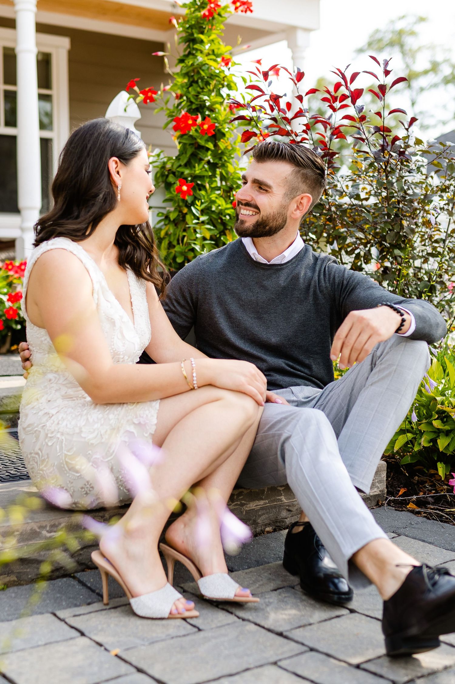 A couple sits together on stone steps with red flowers and greenery in the background.