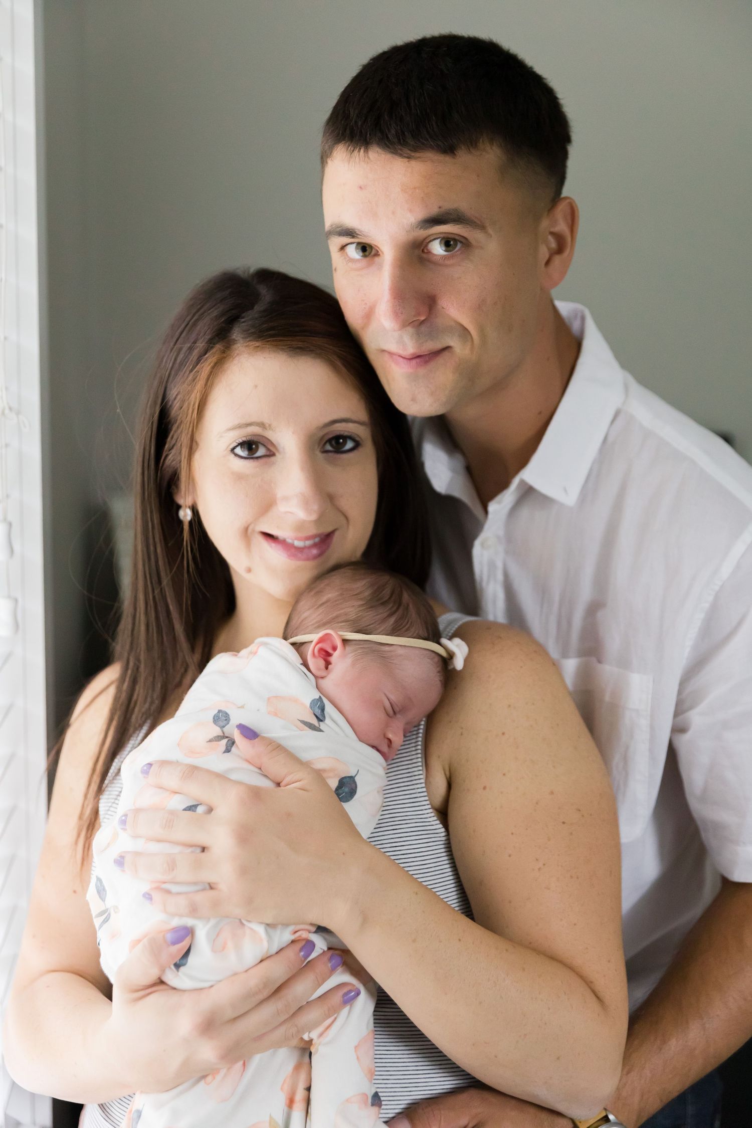 Parents pose with newborn baby by window in natural light.