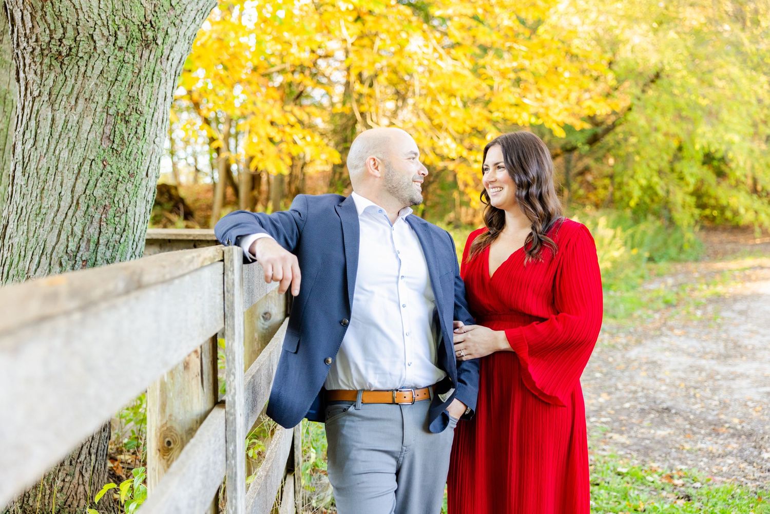 Couple in red dress and navy suit lean against rustic wooden fence during autumn photo session.
