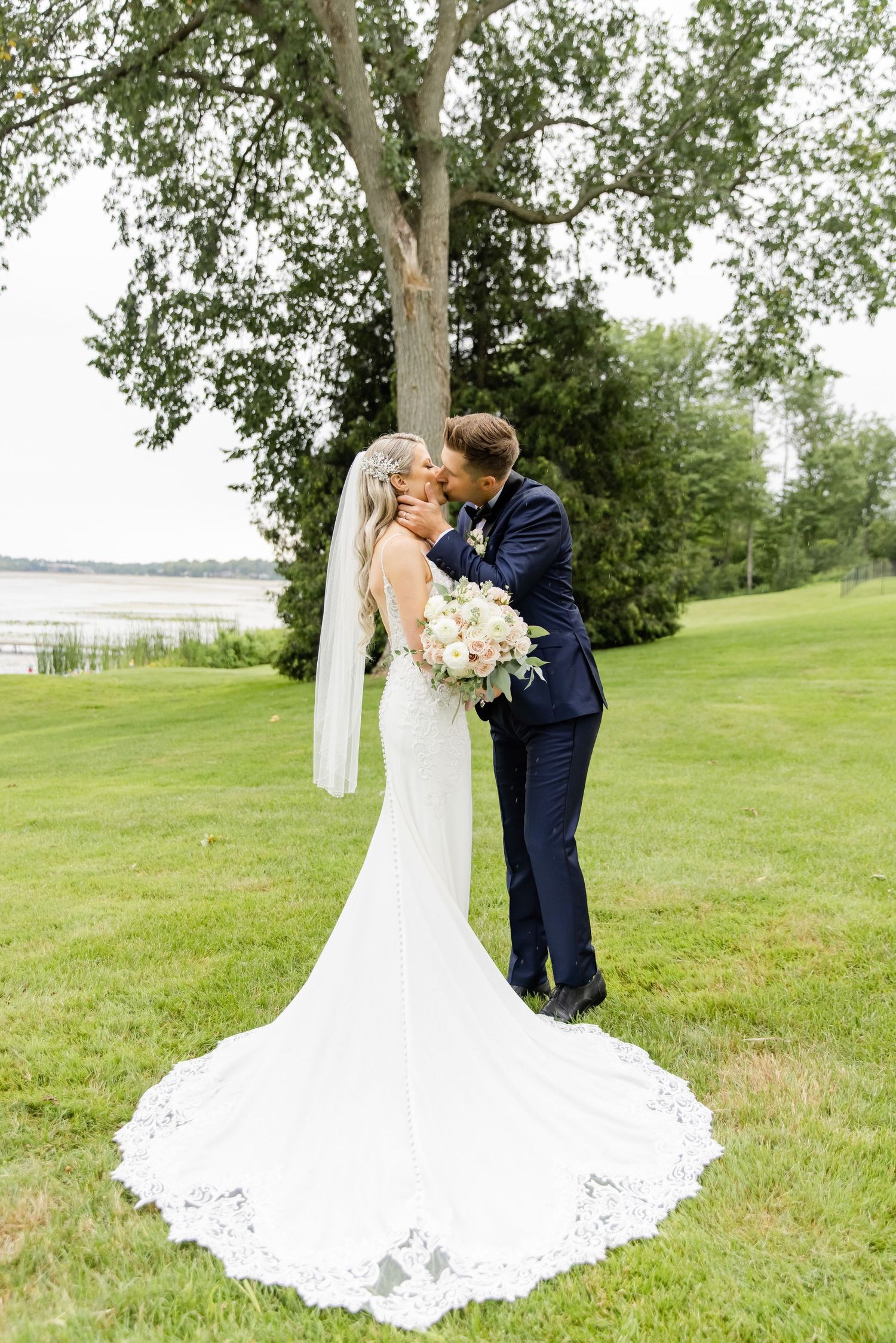 Newlyweds share intimate moment under tree with bride's dramatic train flowing on green grass.