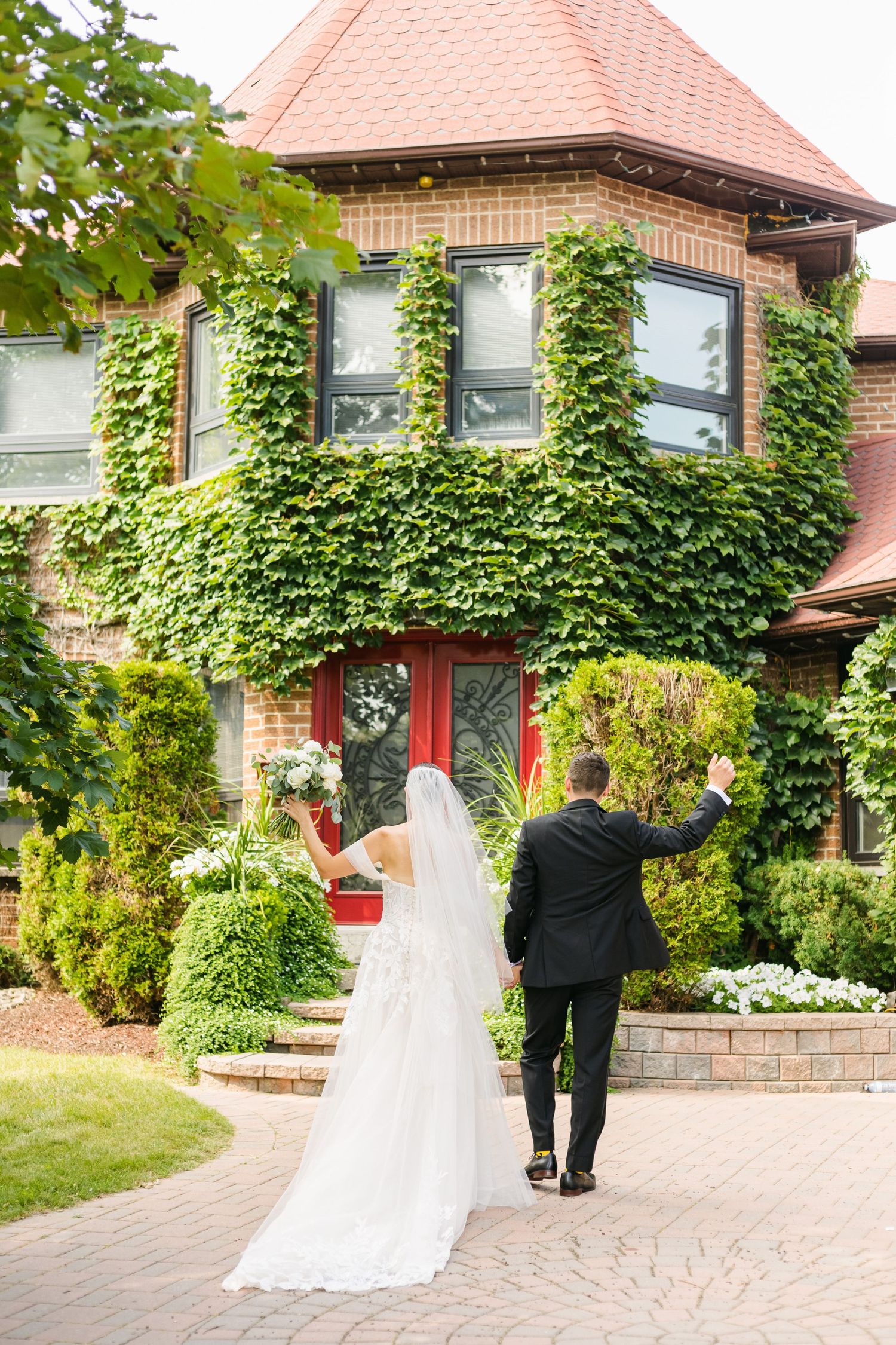Wedding couple walking towards ivy-covered brick house with distinctive red door.