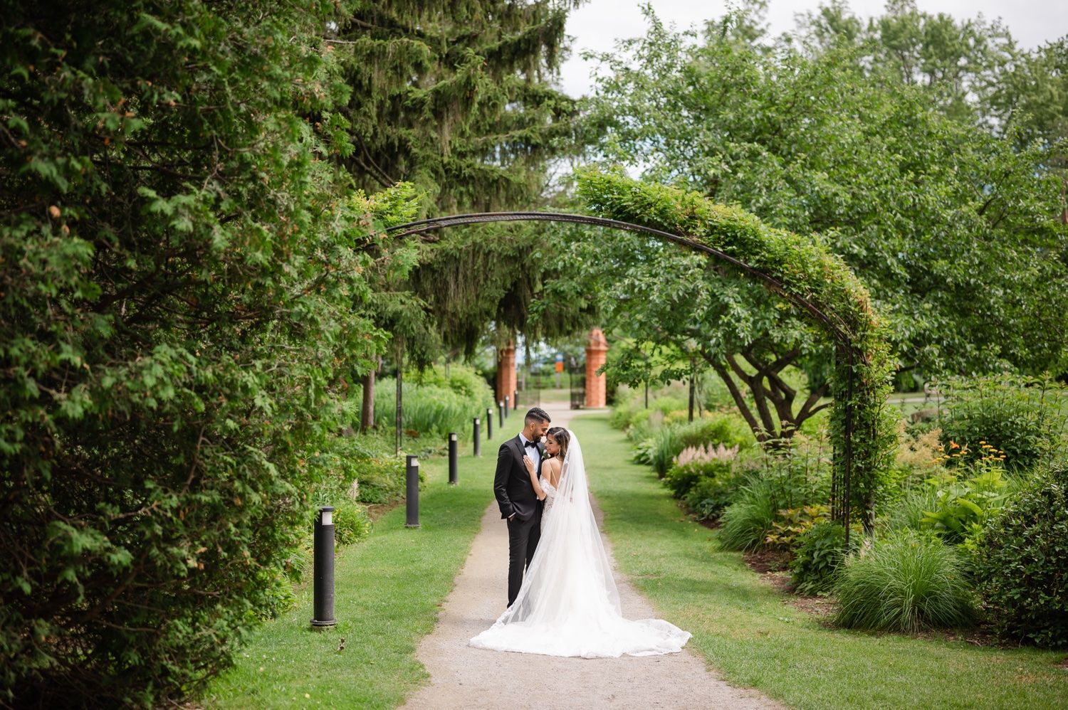 A couple walks along a garden path under a lush green archway on their wedding day.