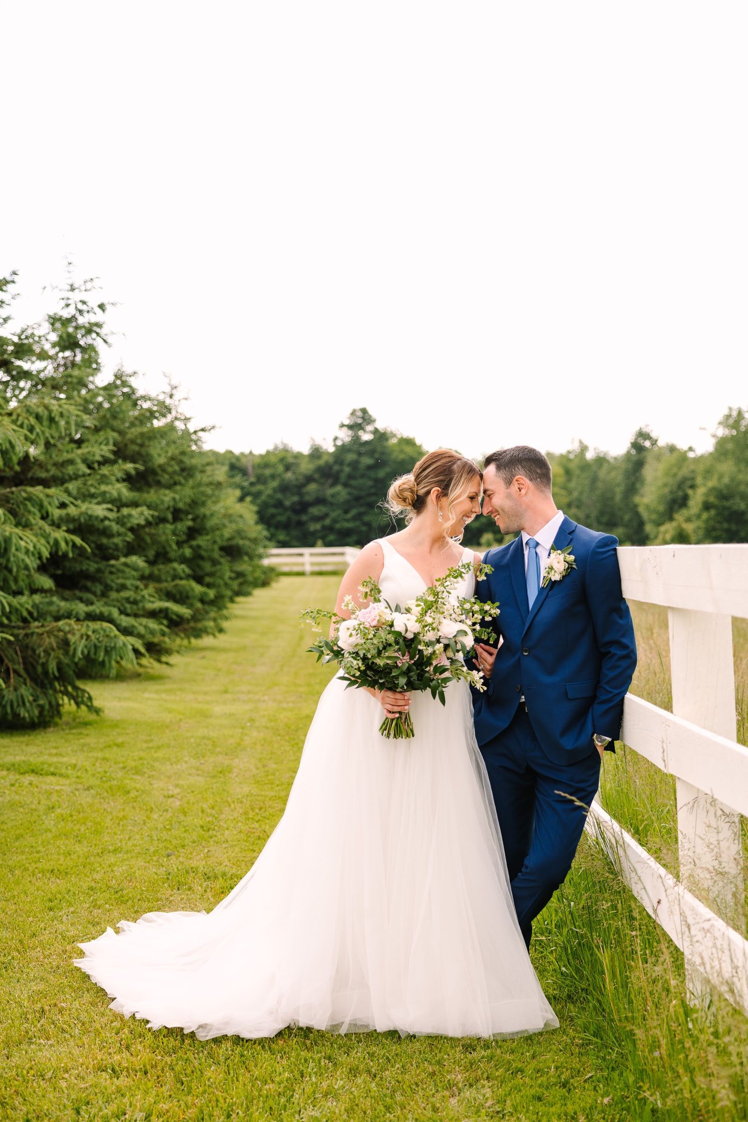 Wedding couple shares intimate moment beside white fence and evergreen trees.
