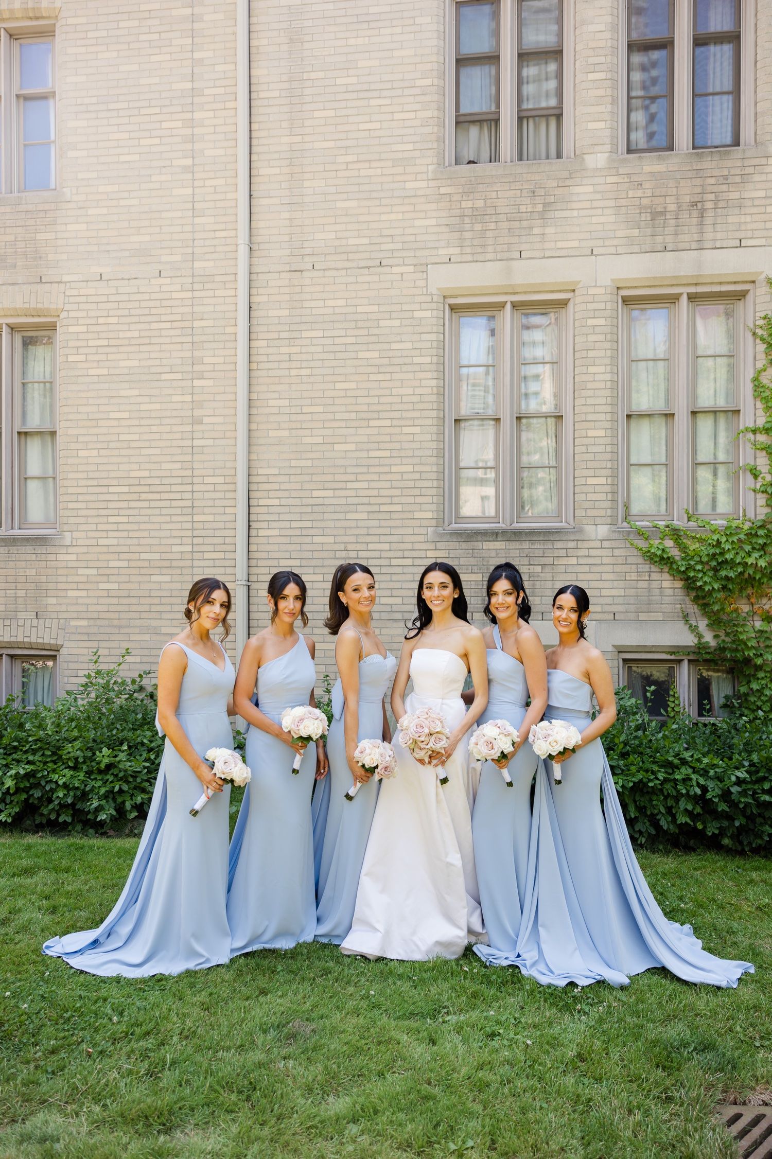 Bridesmaids in flowing powder blue gowns gather around bride in white against brick backdrop.