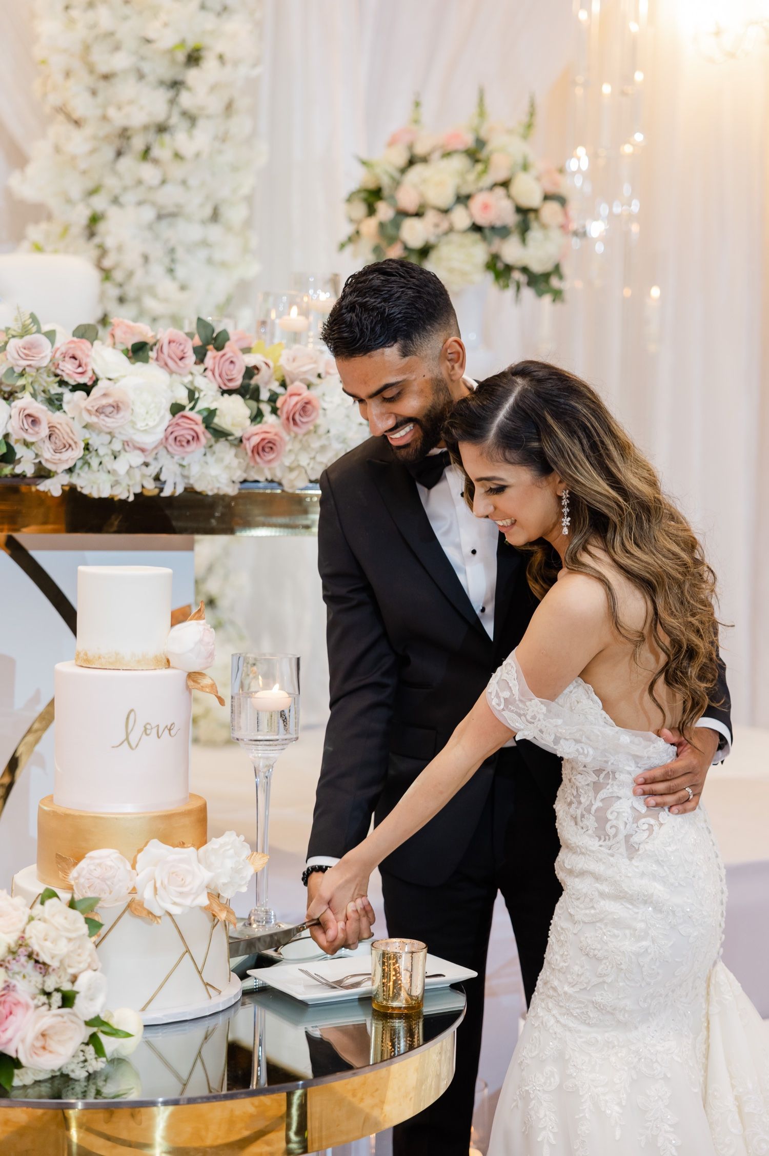 A couple cutting their elegant white and gold wedding cake adorned with roses at their reception celebration.