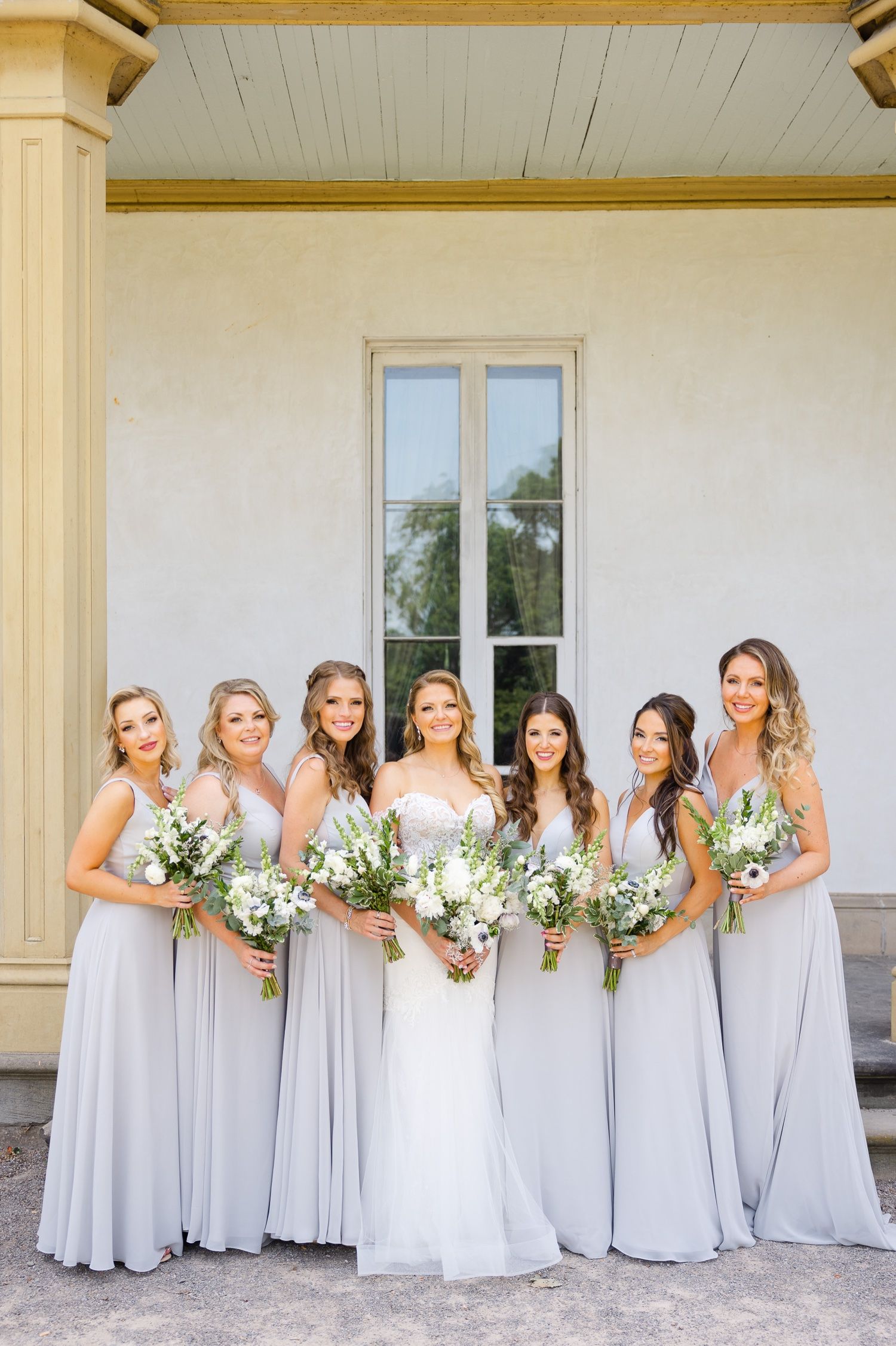 Bridal party in light gray dresses holding white floral bouquets pose by classic architecture.