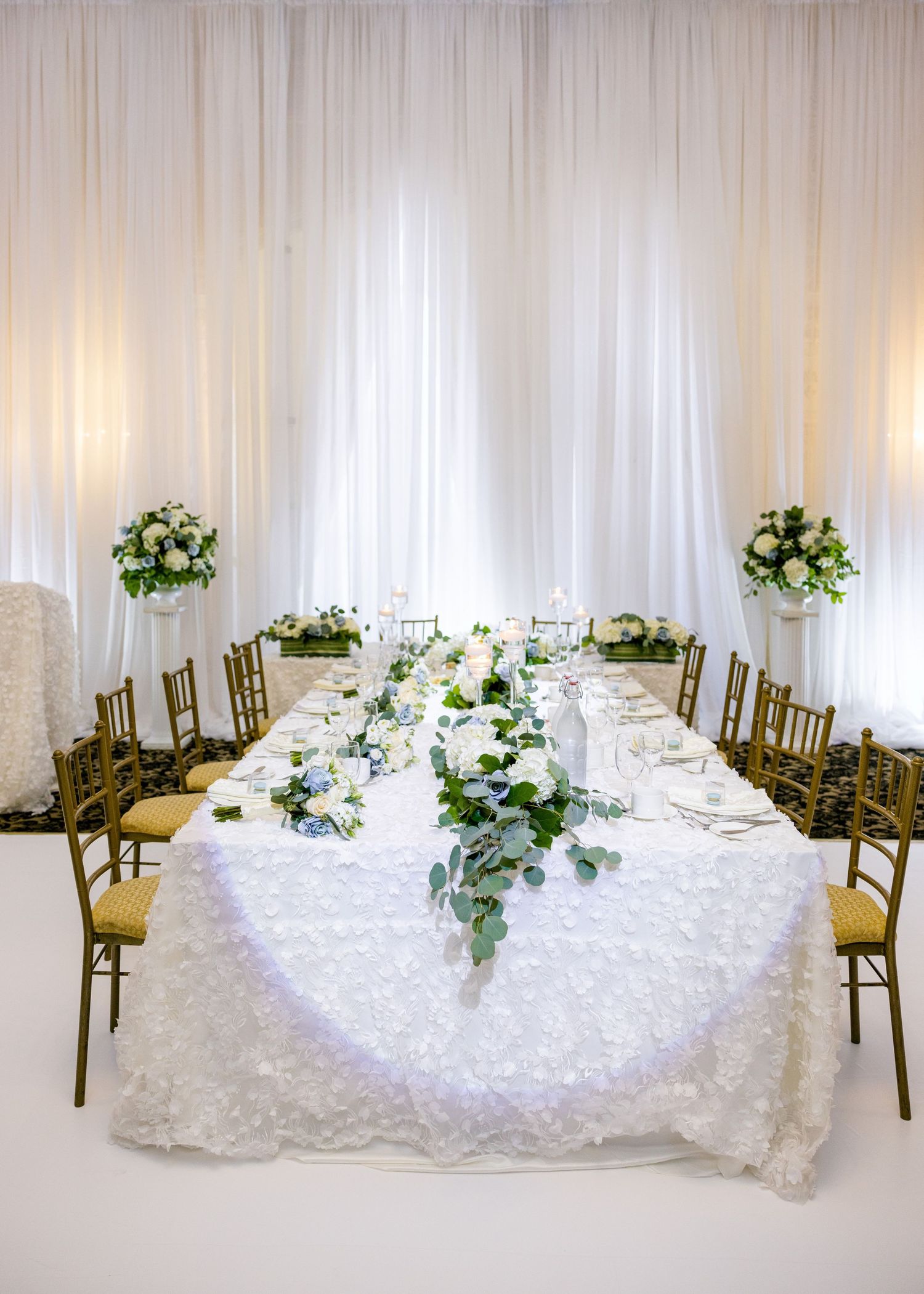 Long banquet table with white lace tablecloth, gold chairs and floral centerpieces against white draping.