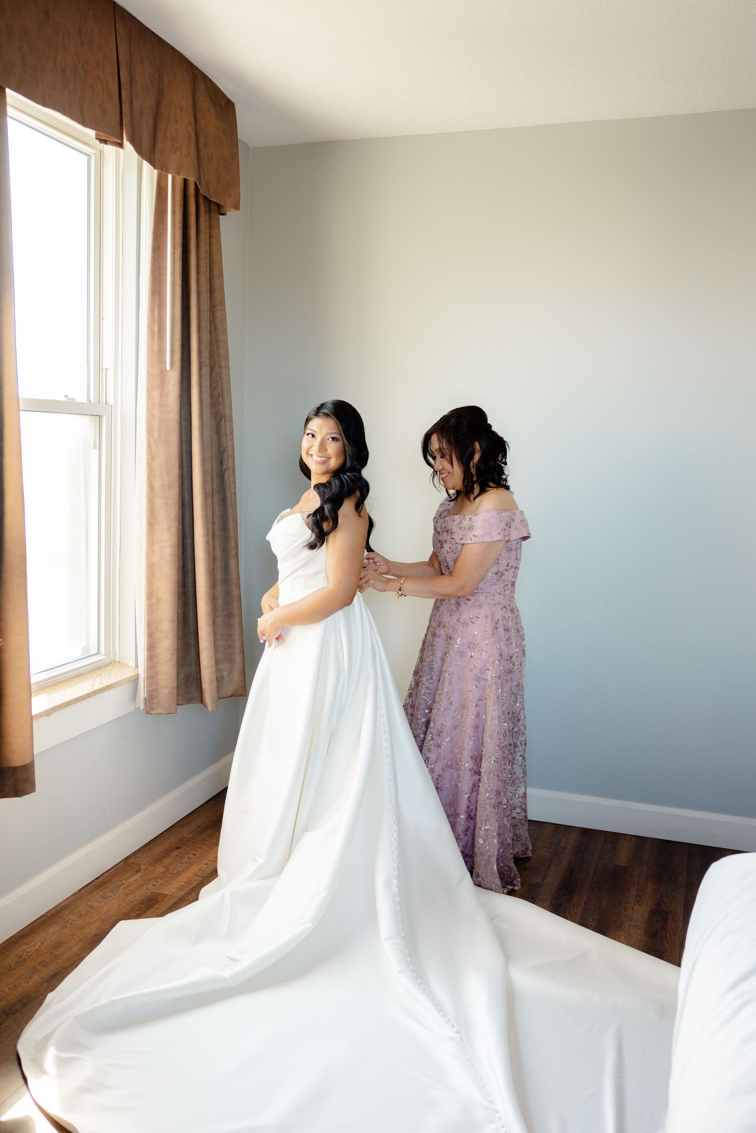 A heartwarming moment as a person in a lavender dress helps adjust a stunning white wedding gown in a sunlit room.