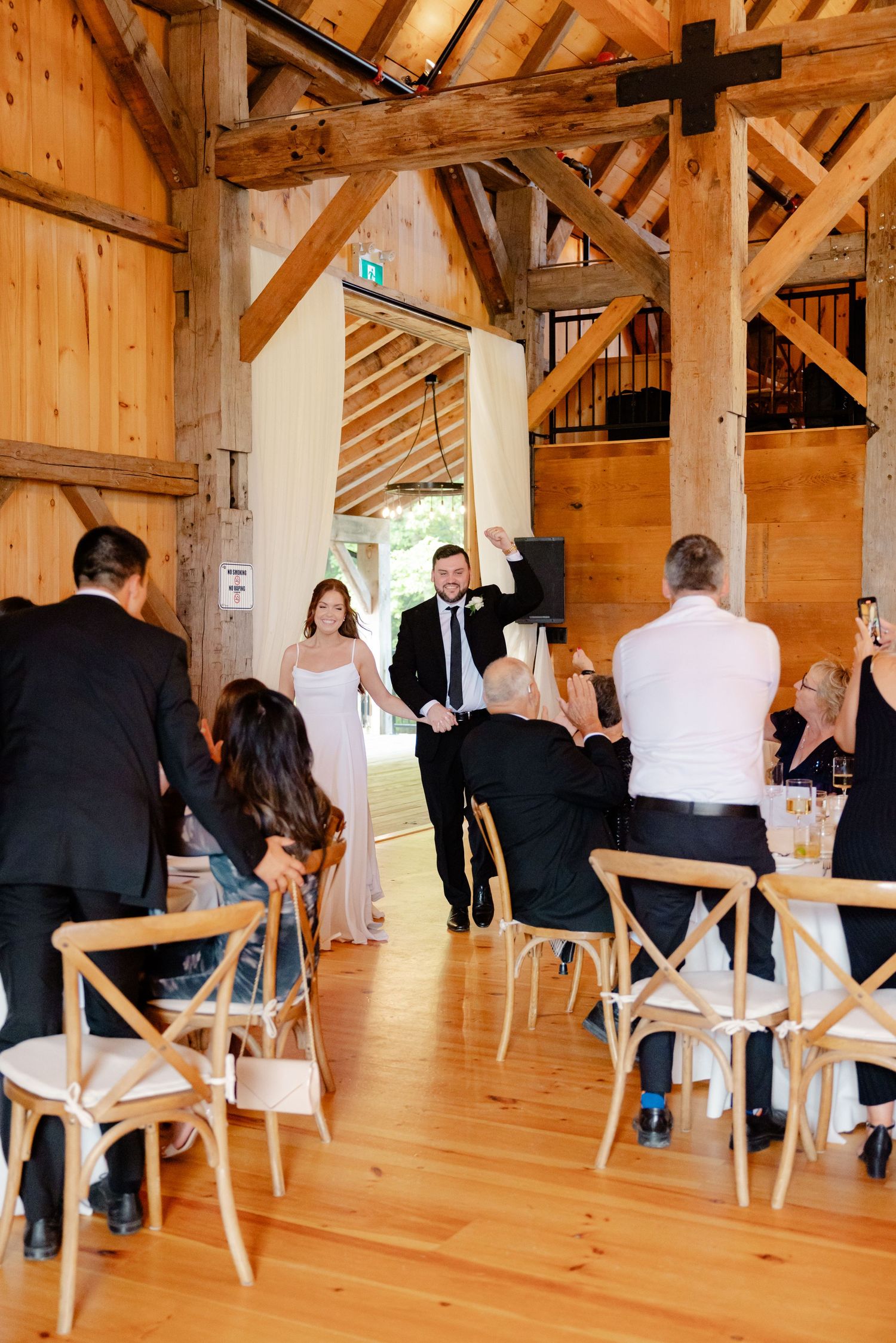 A wedding ceremony taking place in a rustic wooden barn venue with crossback chairs and exposed beam architecture.