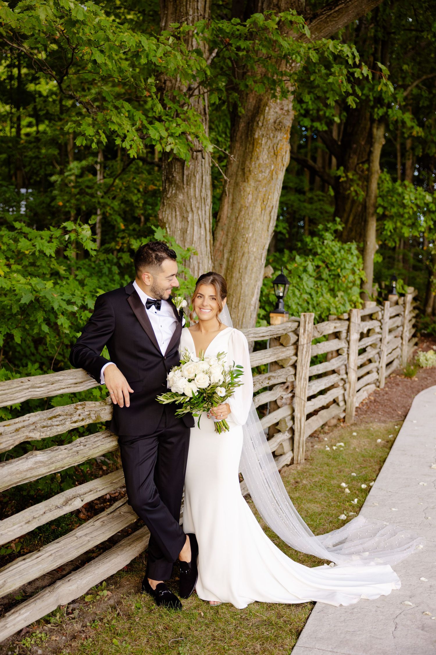 An elegant wedding portrait with the couple posed by a wooden fence surrounded by greenery.