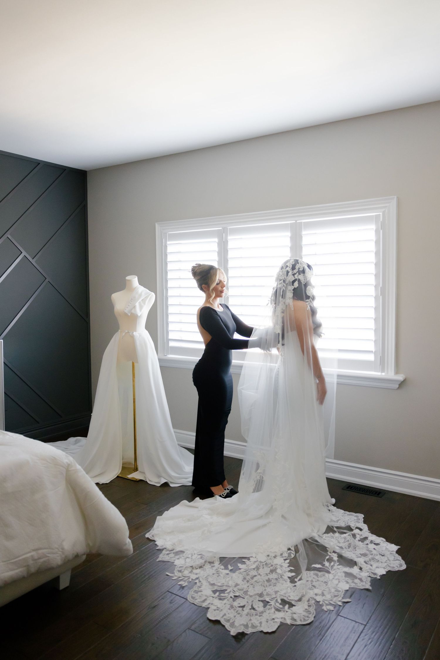 A wedding dress fitting scene with stylist adjusting a long lace-trimmed veil in natural window light.