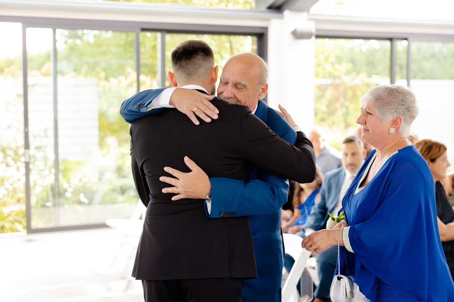 Emotional embrace between wedding attendees during a ceremony in a light-filled venue.