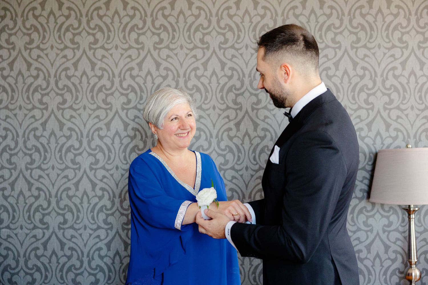A woman in a blue dress shares a tender moment with someone in a black tuxedo against a grey wall.