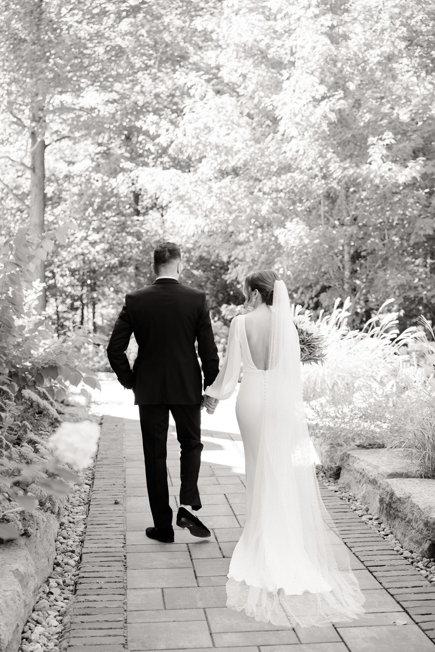 Black and white photo of newlyweds walking down a brick-lined garden pathway.