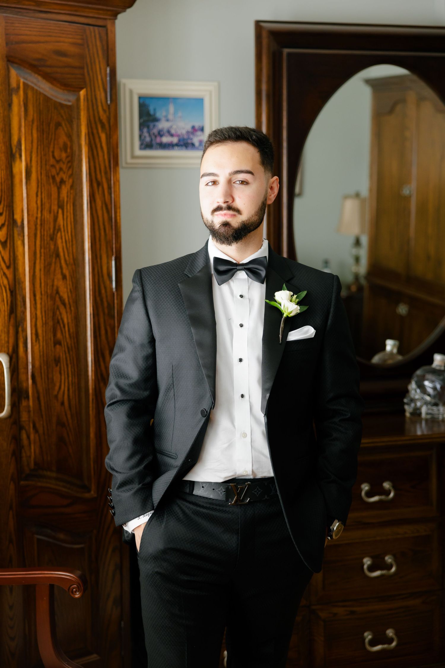 A groom in formal attire poses with one hand in pocket against wooden furniture background.