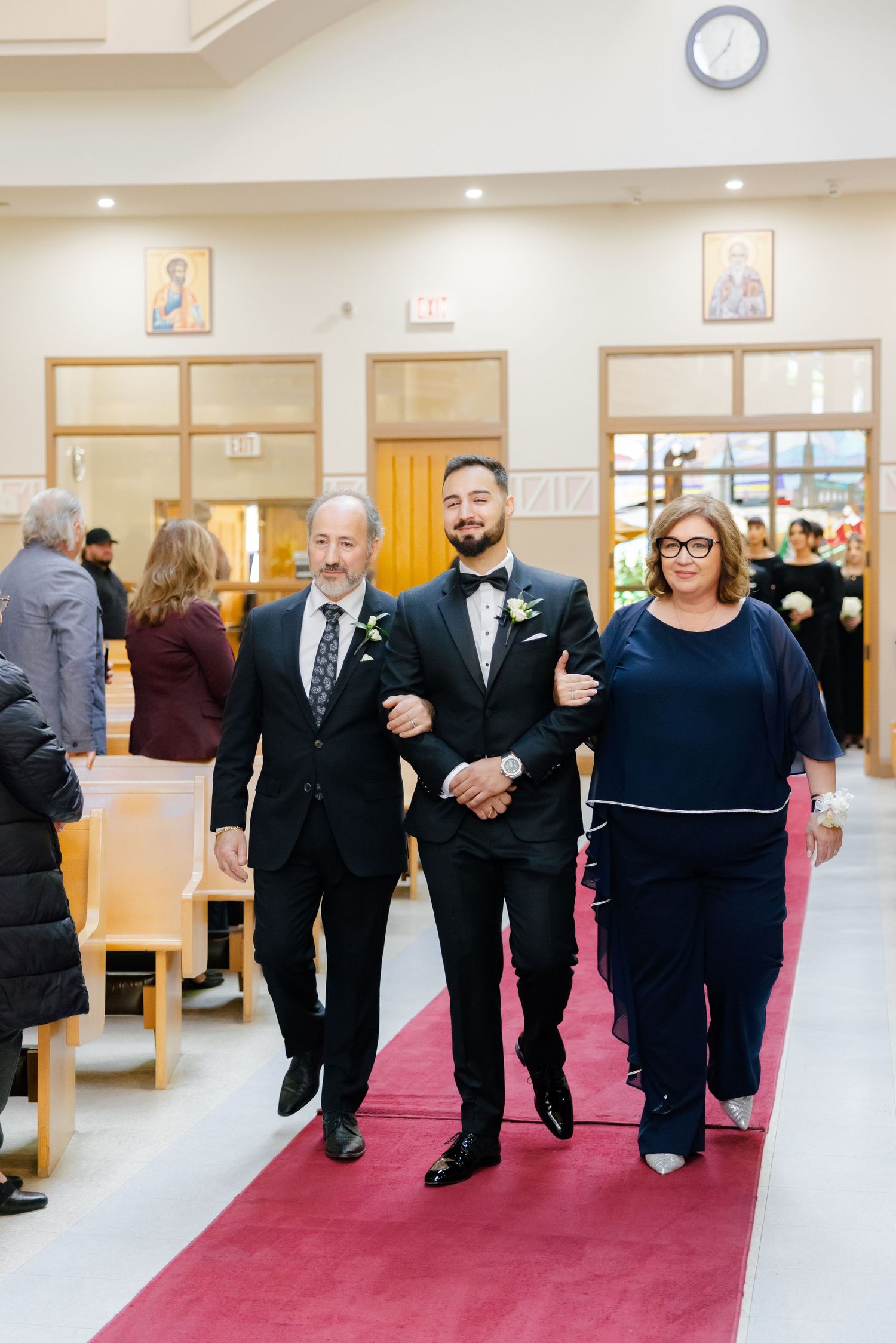 Wedding party members walking arm-in-arm down red carpet aisle in church setting.