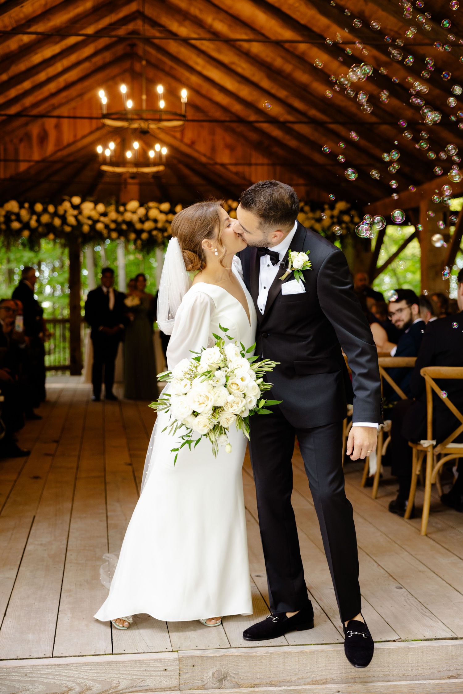 Newlyweds share a romantic moment under wooden beams decorated with string lights at rustic barn wedding venue.