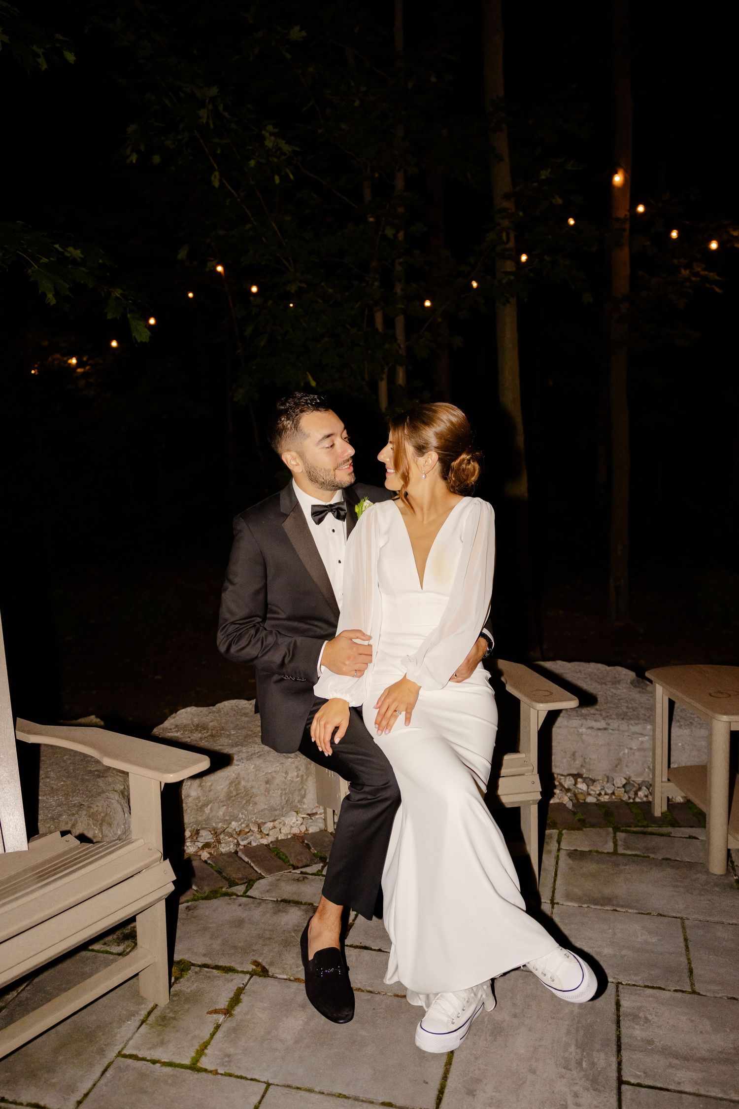 Couple shares intimate moment on stone bench under twinkling lights at nighttime wedding reception.