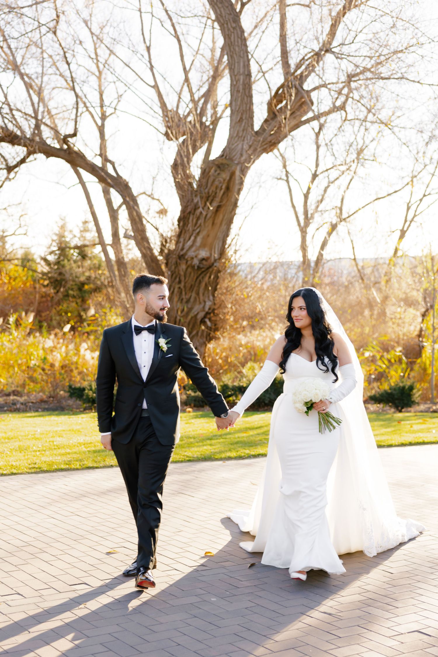 Elegant wedding couple strolling on brick path with autumn trees and golden light.
