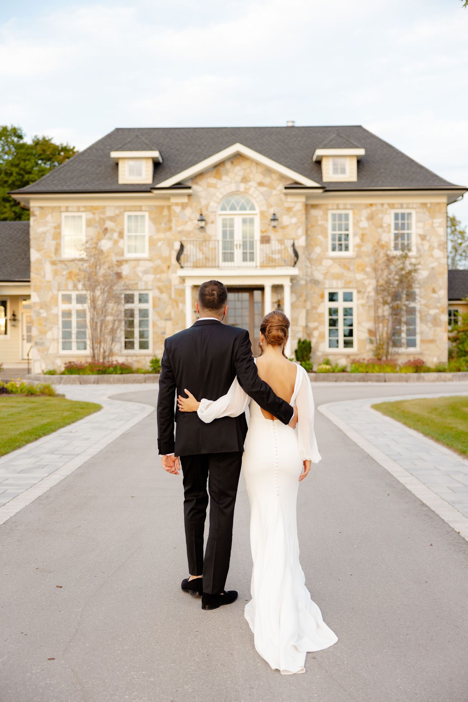 A couple walks arm in arm towards a grand stone estate house on their wedding day.