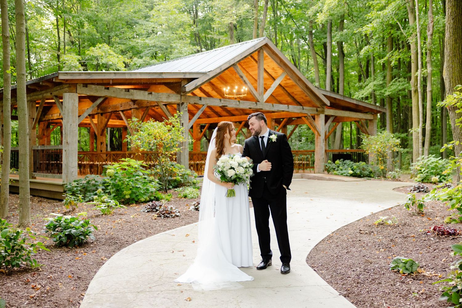 Wedding couple stands on winding path in front of timber-frame pavilion in wooded setting.