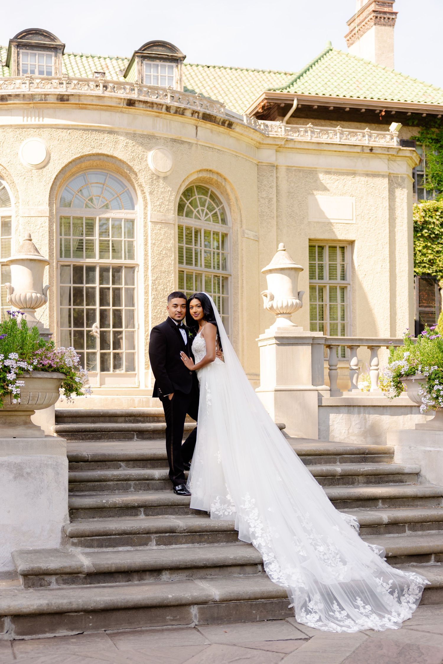 A wedding couple poses on grand stone steps in front of an elegant cream-colored mansion with arched windows.