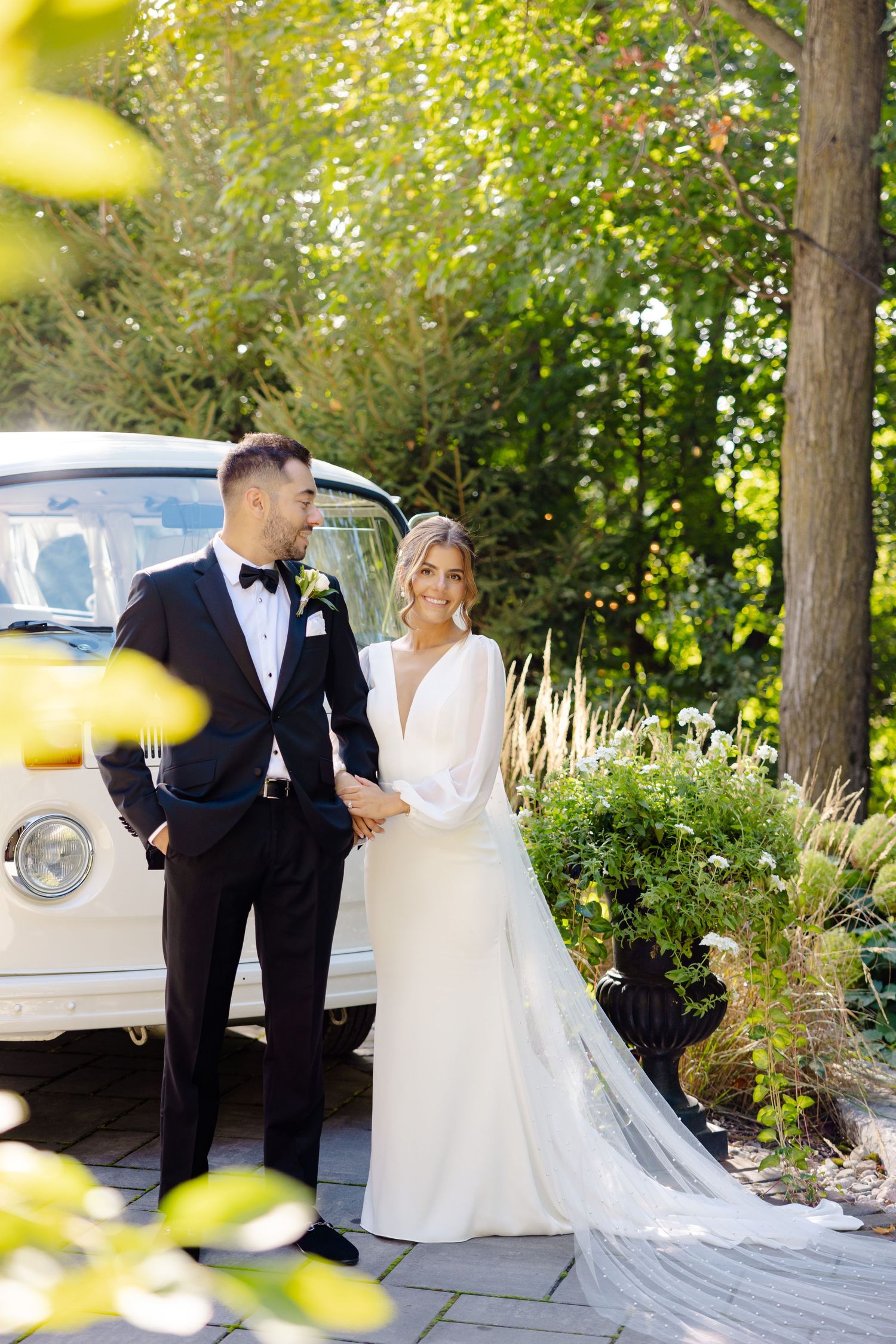 Elegant wedding couple poses by vintage vehicle with sunlit garden and stone path backdrop.