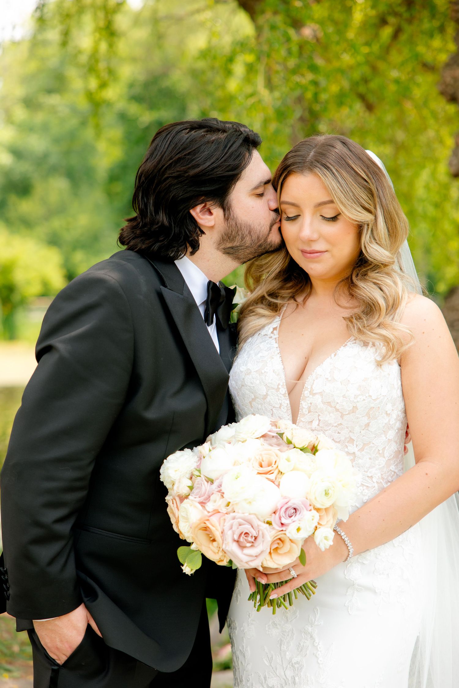 Romantic moment between newlyweds with soft pink bouquet under garden willow tree.