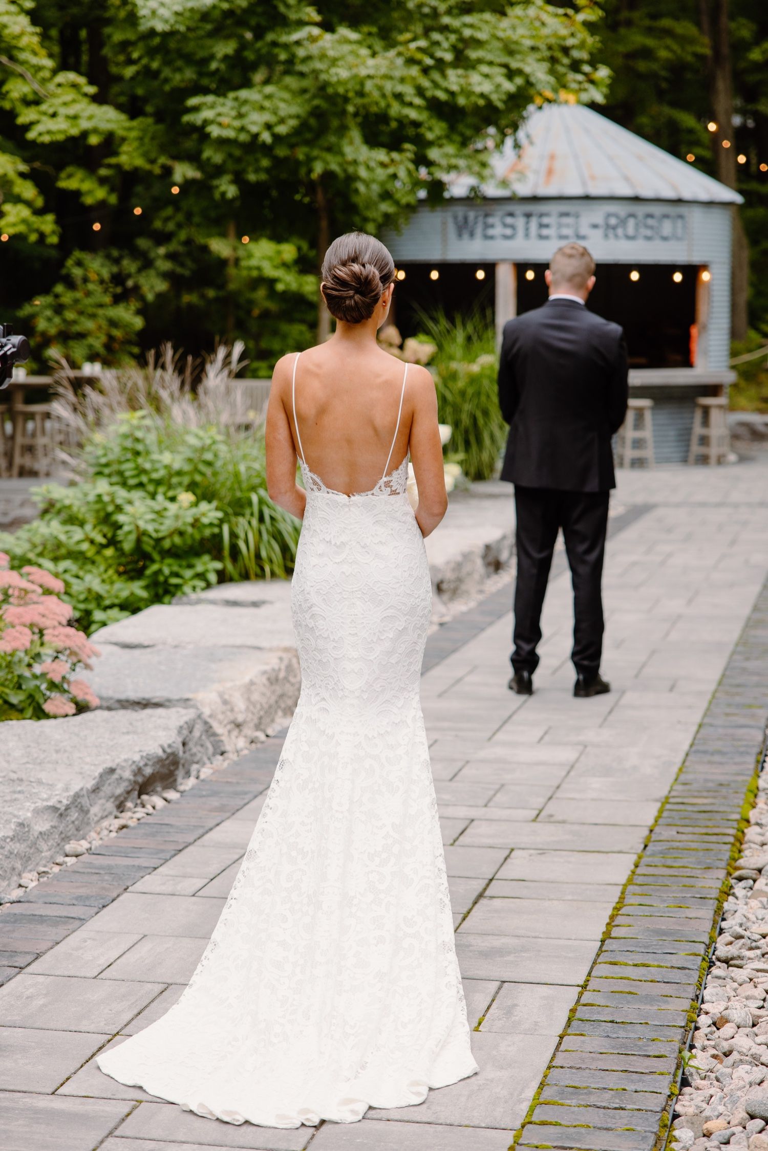 Back view of bride in fitted lace gown with low back detail walking on stone path.