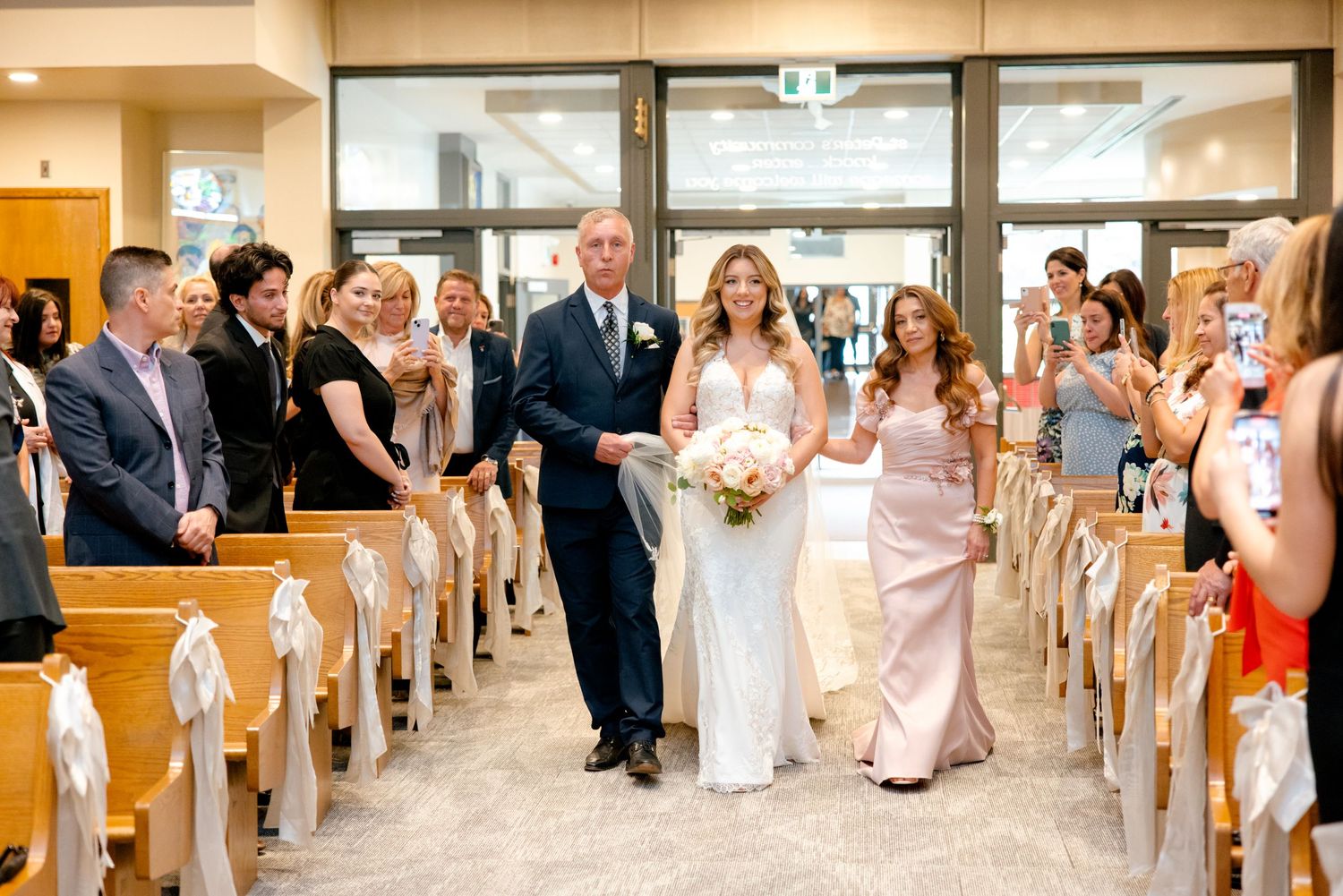 Wedding ceremony processional in bright church hall with guests watching from decorated pews.
