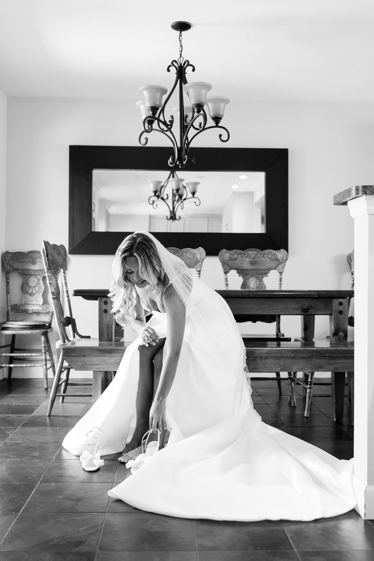 An elegant bride adjusts her shoes in a dining room with a large mirror and chandelier.