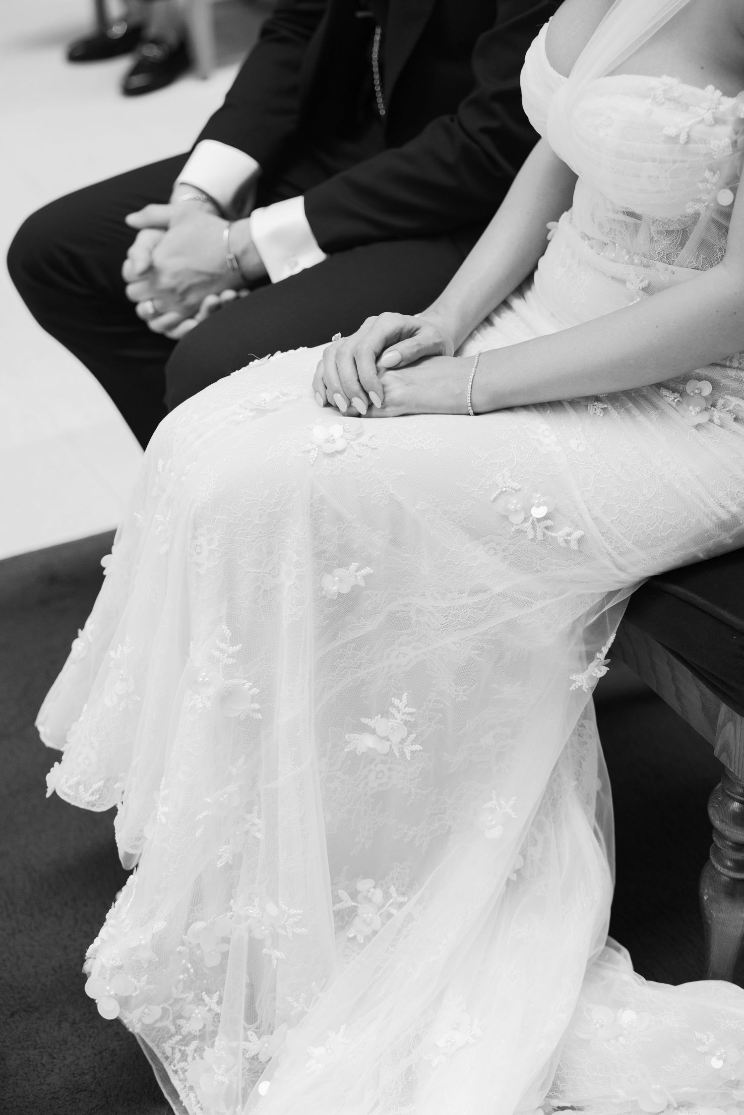 A close-up black and white detail of clasped hands and wedding dress lace during the ceremony.