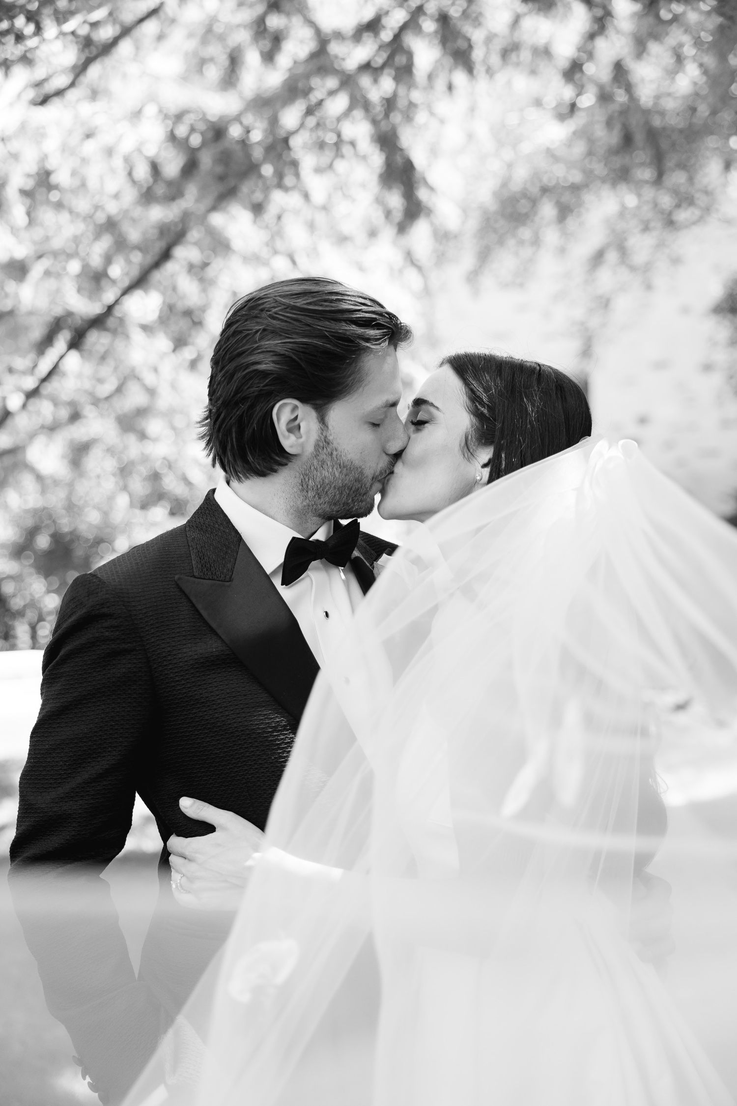 A romantic black and white wedding photograph capturing a tender kiss between newlyweds with a flowing bridal veil.