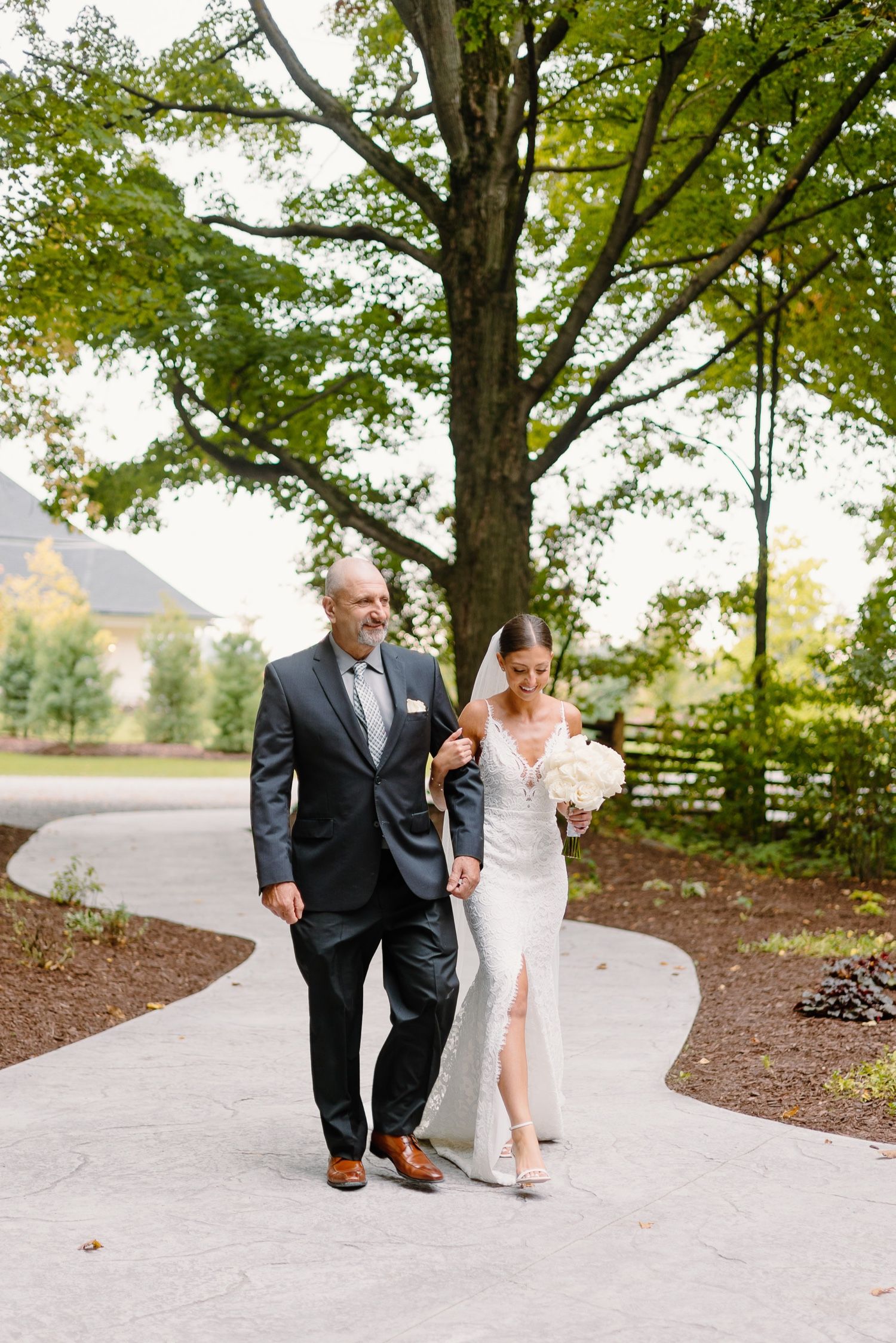 Elegant wedding couple walking down curved pathway with trees and building in background.