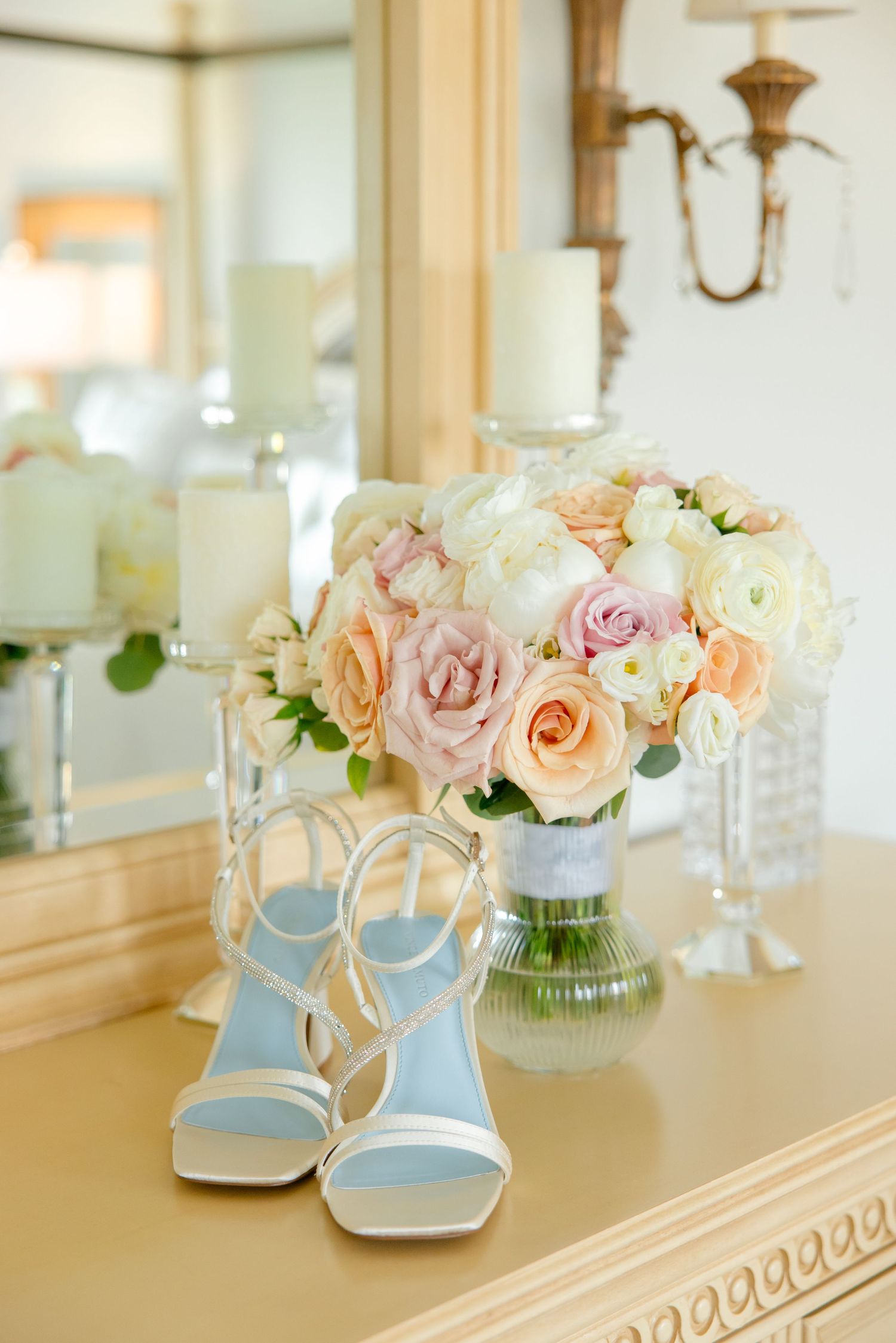 Elegant wedding details with pale pink and peach roses in a vase next to light blue strappy heels on a vanity.