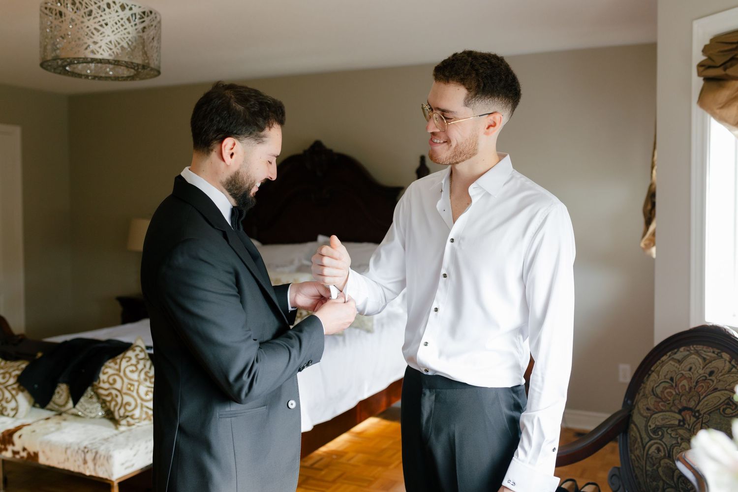 Two people helping each other get dressed in formal attire in a bedroom setting.