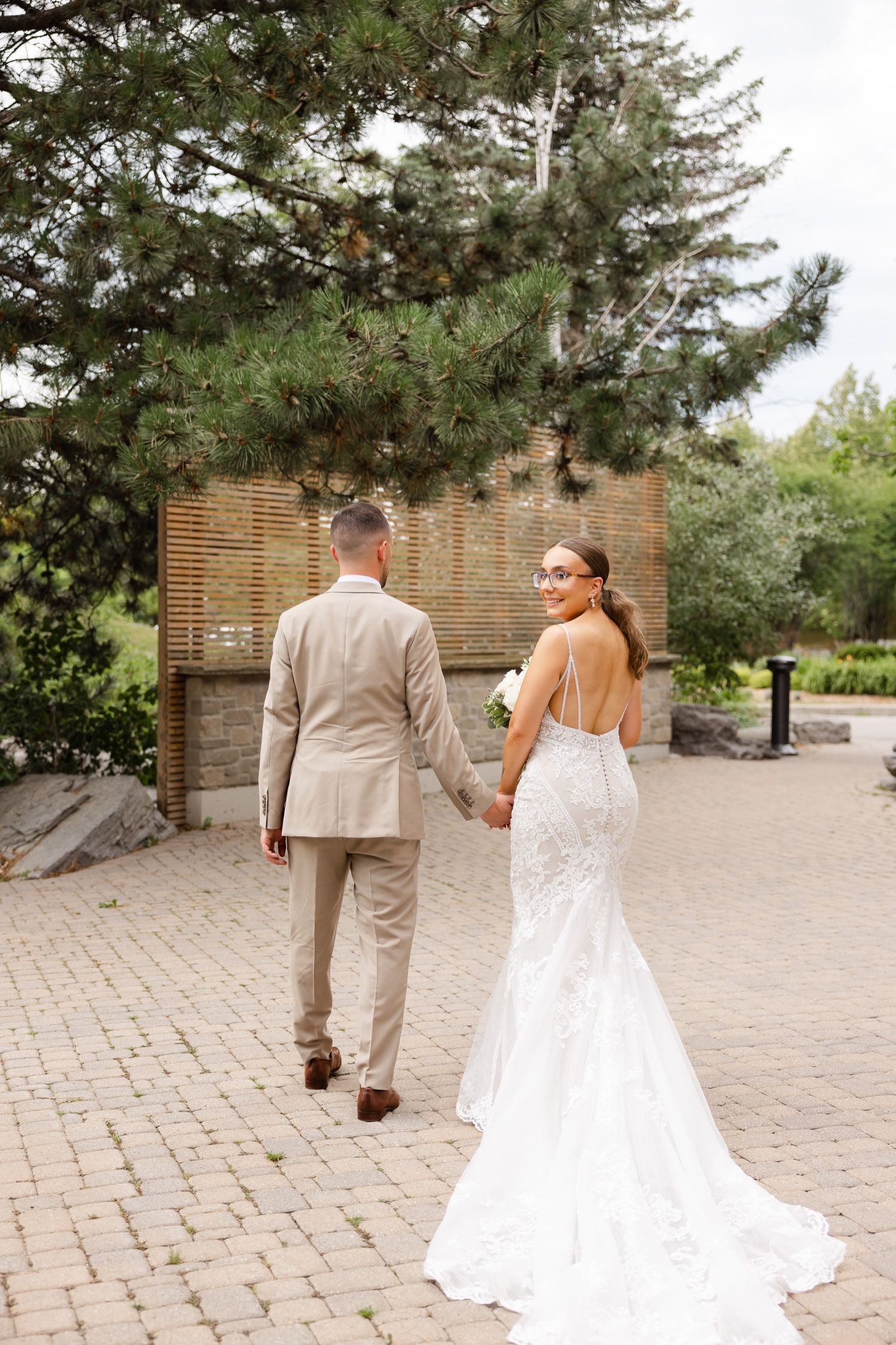 A wedding couple walking on a stone path with lush pine trees and wood paneling in the background.